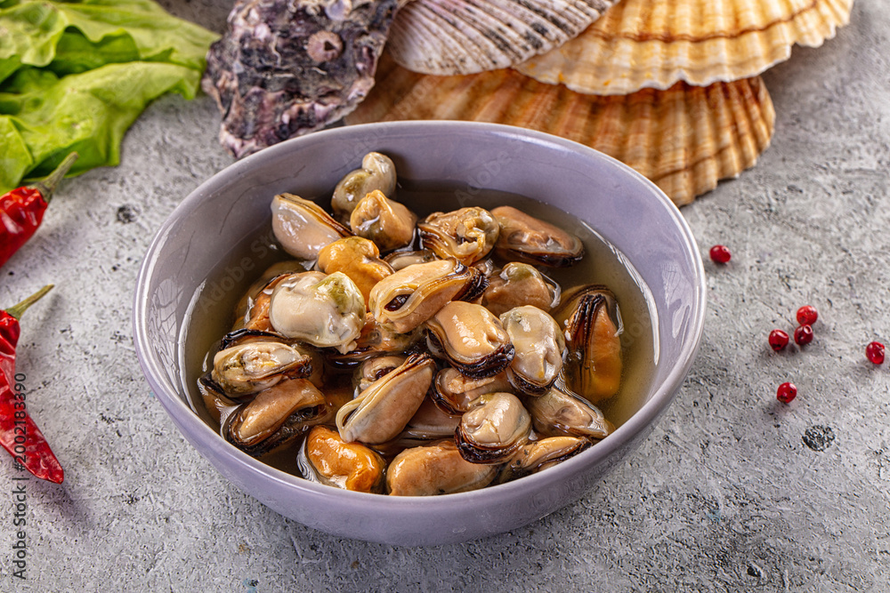 Fototapeta premium Marinated mussels in a bowl with chili peppers, lettuce, and seashells on a rustic grey background.