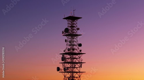 Tall telecommunications tower silhouetted against a stunning gradient sunset sky transitioning from purple to orange. Multiple satellite dishes and antennas with blinking red warning lights visible.