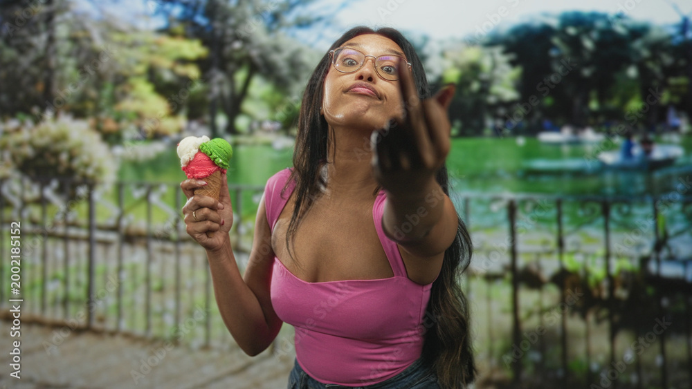 Fototapeta premium Woman in pink top and glasses holding a multicolored ice cream cone flips middle finger by a lakeside fence in street; sassy defiance.