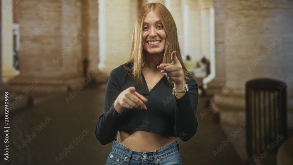 Obraz premium Redhead woman pointing finger and showing bare midriff beneath a black cropped top in a stone building archway with passerby blur; youthful confidence.