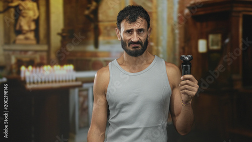 Canvastavla Man holding electric shaver with beard visible and grimace beside votive candles at a church building; unease