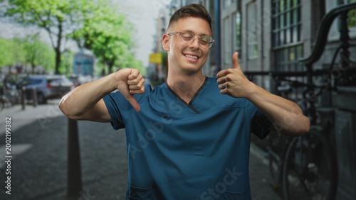 Φωτογραφία Man doctor in blue medical scrubs smiling with glasses showing thumbs up and thumbs down while standing on a street; ambivalence choice
