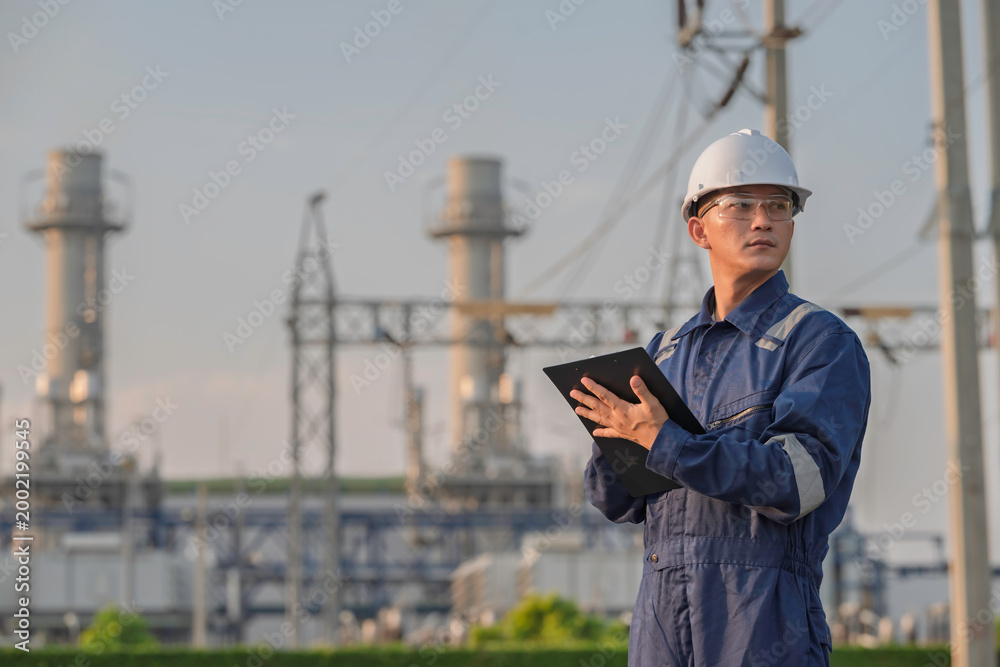 Fototapeta premium Asian male electrical engineer holding digital tablet standing at power plant station. Professional energy industry technician working on infrastructure maintenance and renewable resource project.