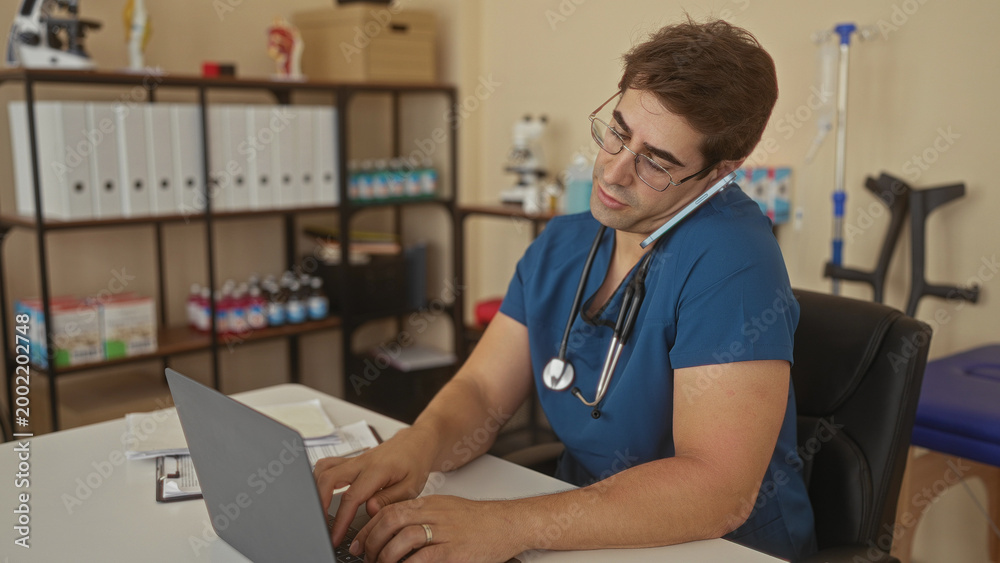 Fototapeta premium Man doctor with stethoscope wearing blue scrubs and glasses holds phone to ear and types on laptop in building; focused care.