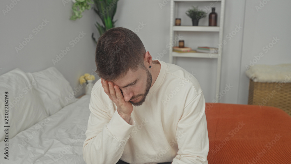Fototapeta premium Man sitting on bed with hand on face and head in hand gesture inside building; quiet reflection sadness.