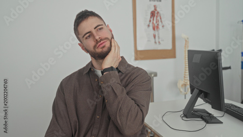 Foto Man touching jaw with hand and wincing, hand on cheek and eyes closed, seated at desk by monitor and anatomy poster in building clinic; pain concern