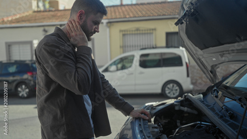Cuadro en lienzo Man with hand on neck examining open car hood on street near parked vans; concern repair troubleshooting