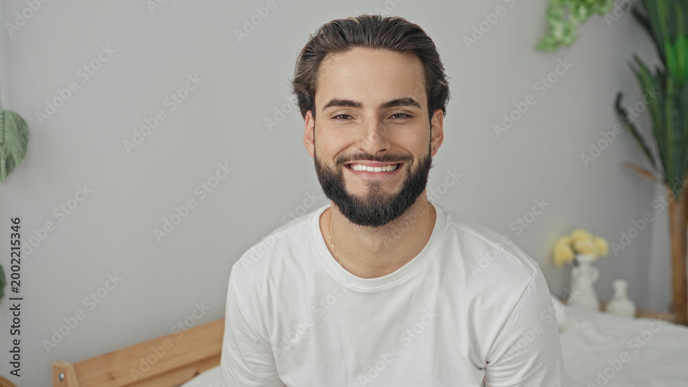 Obraz premium Man on bed smiling, face and beard visible, wearing white tshirt and gold chain, facing camera in studio; relaxed contentment.