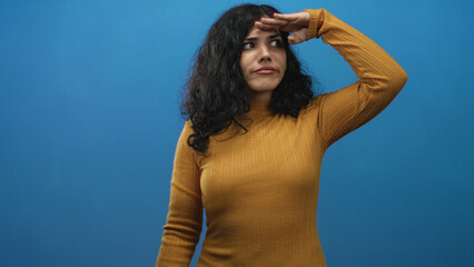 Young arab woman shielding eyes with hand while scanning the distance in a blue studio wearing a mustard sweater and curly hair; curiosity searching.