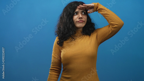 Fotografija Young arab woman shielding eyes with hand while scanning the distance in a blue studio wearing a mustard sweater and curly hair; curiosity searching