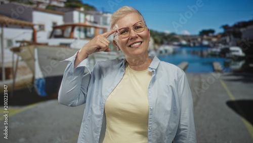 Slika na platnu Woman points finger to head on a seaside street with boats and dock visible; joyful travel memory recollection