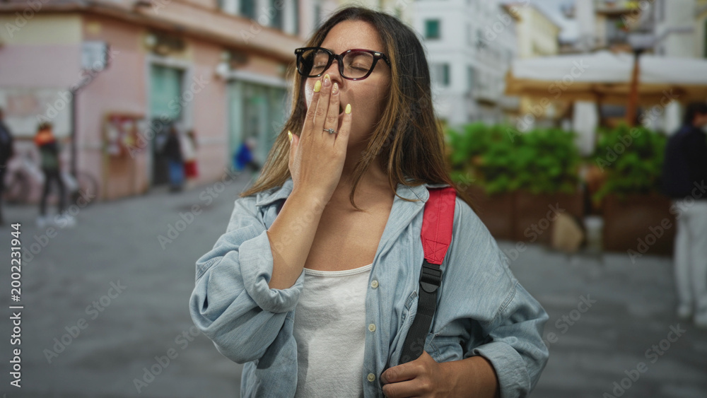 Obraz premium Woman covering mouth with hand and eyes closed while yawning on a city street with backpack visible; sleepiness.