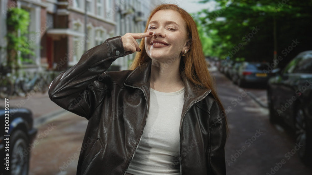 Fototapeta premium Woman pointing finger to nose on a street in front of parked cars and buildings, wearing leather jacket and smiling broadly; confidence openness.