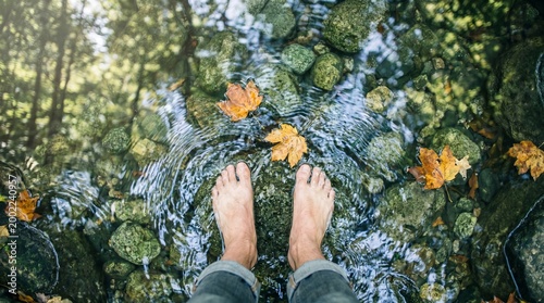 Person wading barefoot in shallow stream surrounded by autumn leaves