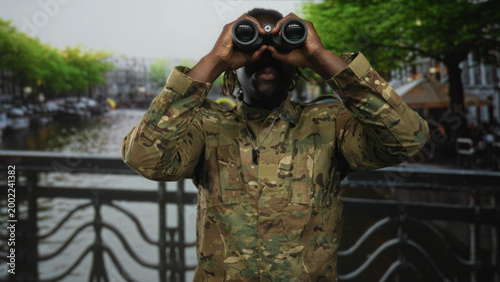 фотографія Man soldier in camouflage holds binoculars to eyes on a canal bridge in amsterdam street; duty vigilance