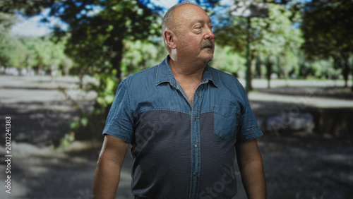 Man standing with bare forearm visible, looking to the right with a slight head turn in a sunlit park; quiet contemplation.