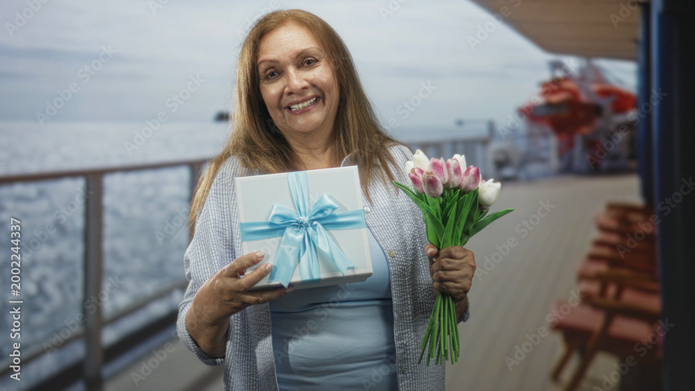 Fototapeta premium Woman holding a ribboned gift and a tulip bouquet on a cruise deck smiling gently; celebration joy gratitude.
