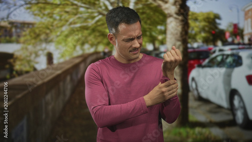 Photos Young hispanic man clutching wrist with pained grimace on a street beside parked cars and stone railing; pain recovery