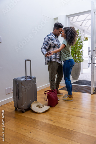 Couple standing holding hands and leaning heads in foyer near gray suitcase, red handbag, straw hat