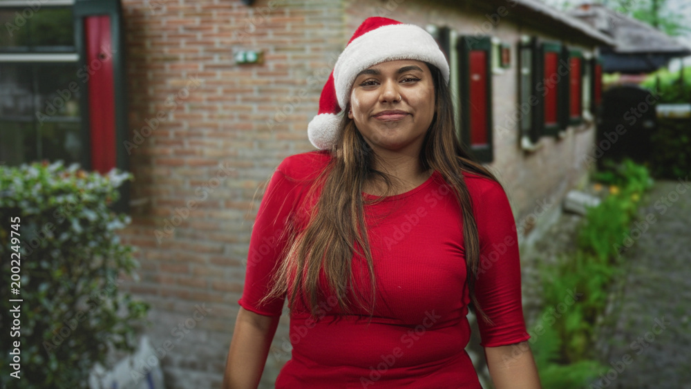 Fototapeta premium Woman wearing santa hat smiling and raises fists while swaying in red top in front of building with brick wall and window shutters; holiday joy.