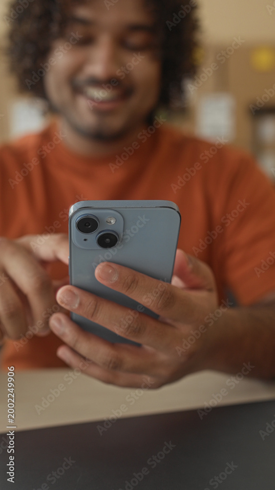 Fototapeta premium Smiling man holding smartphone, finger tapping screen amid cardboard boxes and laptop on desk in building; satisfaction entrepreneurship.
