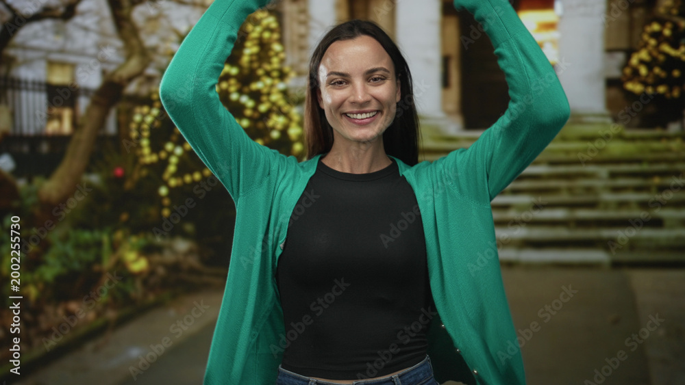 Obraz premium Woman smiling with arms raised and hands visible in front of building steps and twinkling lights near an entrance; joy celebration.