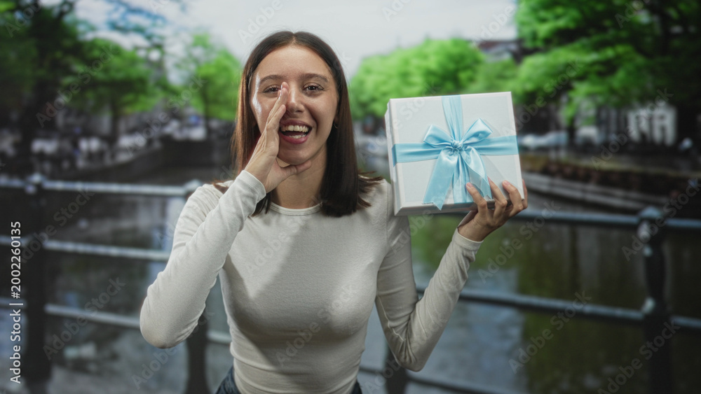 Obraz premium Young woman holding a wrapped gift box with blue ribbon and cupping her hand to mouth on a street by a canal railing, smiling broadly; surprise joy celebration.