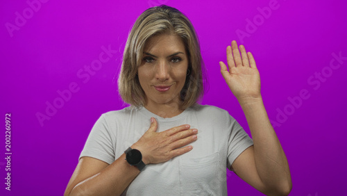 Young blonde woman showing hand on chest and raising hand in a purple studio setting; serious commitment.