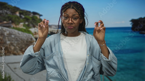 Fotografie Woman wearing glasses and denim shirt holding hands in air quotes gesture at studio seaside set with turquoise ocean backdrop; skepticism irony