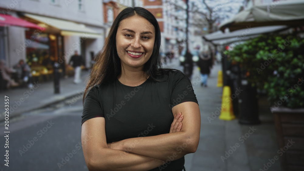 Fototapeta premium Young hispanic woman with crossed arms showing forearms and smiling on a busy city street cafe setting; confidence empowerment.