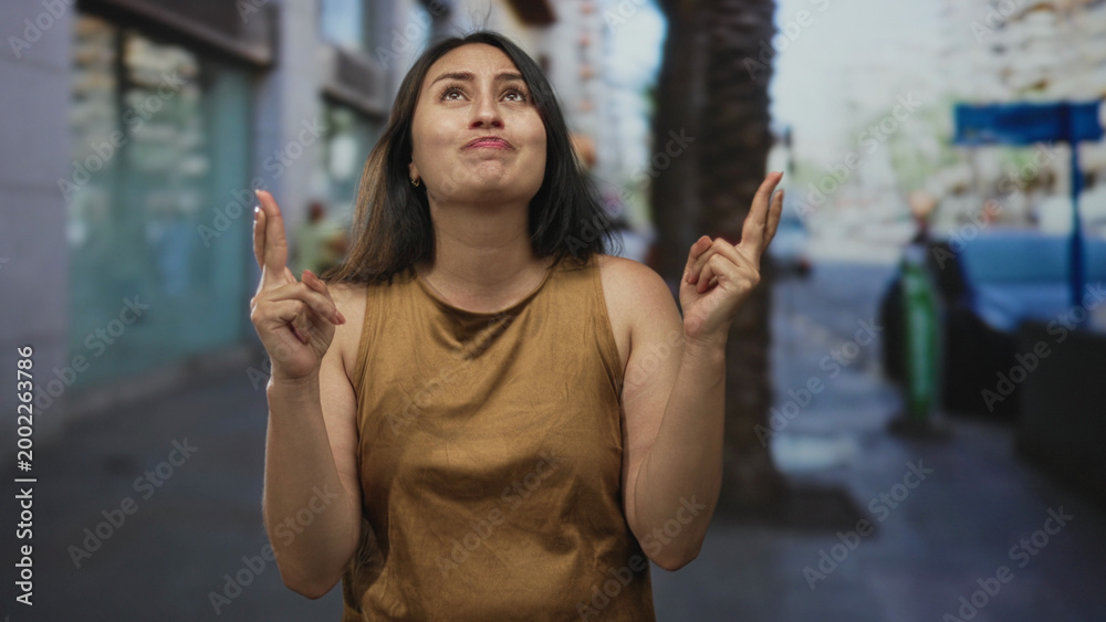 Fototapeta premium Woman with crossed fingers smiling on city street wearing brown sleeveless shirt and small earrings; hopeful wish.