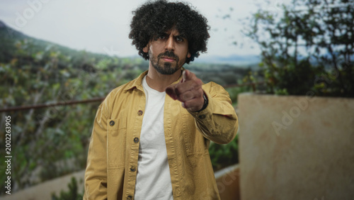 Obraz na plátne Man points finger at camera on building terrace outdoors, curly hair and beard, yellow jacket and white t shirt; accusation confrontation resolve