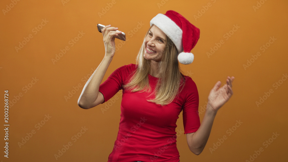 Naklejka premium Blonde woman holding smartphone to mouth singing and smiling in a studio with santa hat and red shirt; joyful celebration.