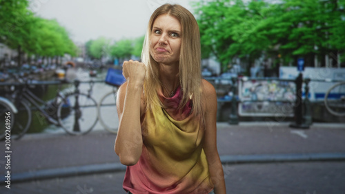 Foto Blonde woman with clenched fist raised, tense arm and angry face, gesturing toward camera beside bicycles by a canal on a street; anger defiance