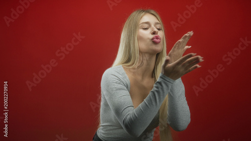 Fotografie Woman blowing a kiss with hands and puckered lips in a studio against a vivid red backdrop captured midgesture; playful affection