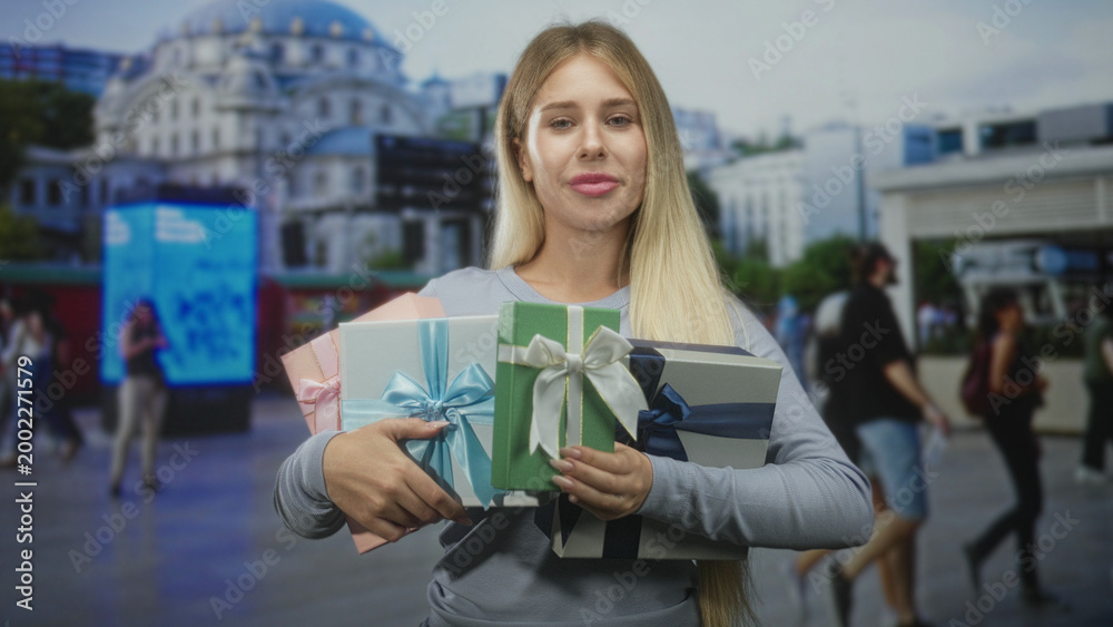 Fototapeta premium Woman holding stacked gift boxes with hands, smiling with long blonde hair and casual sweater on street near building; celebration joy.