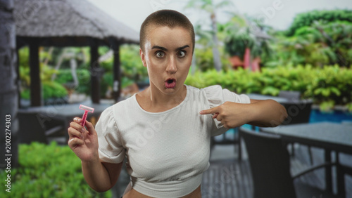 Tableau sur toile Young woman with shaved hair holding a razor points finger to chest showing bare midriff in building with gazebo and pool background; surprise confidence