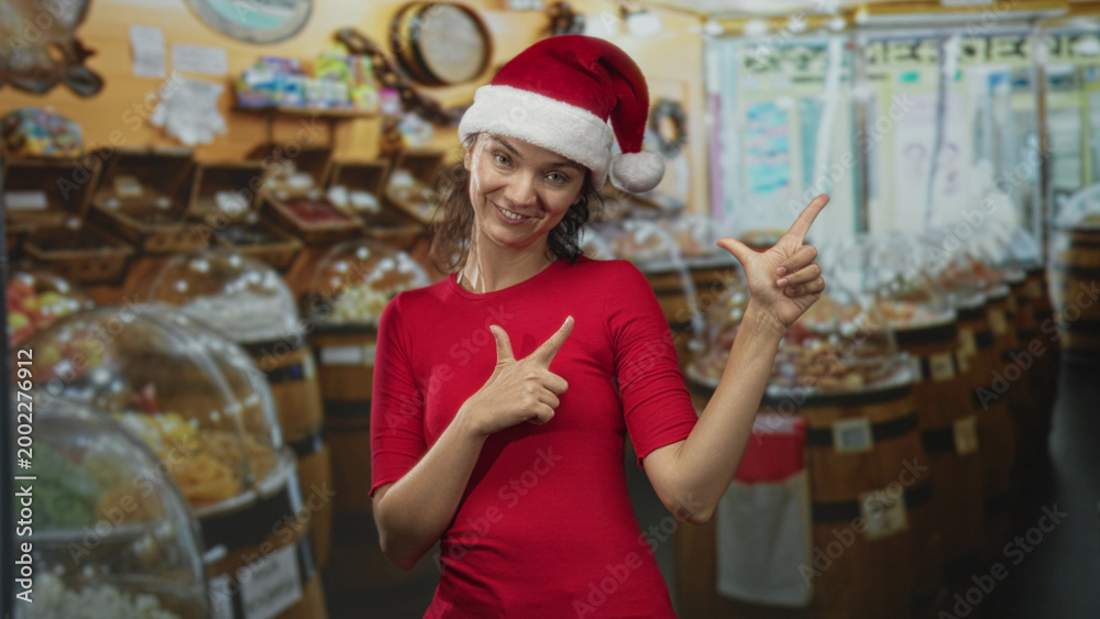Fototapeta premium Woman in santa hat and red shirt points finger to the right and makes a finger gun gesture in a candy shop; holiday cheer festive.