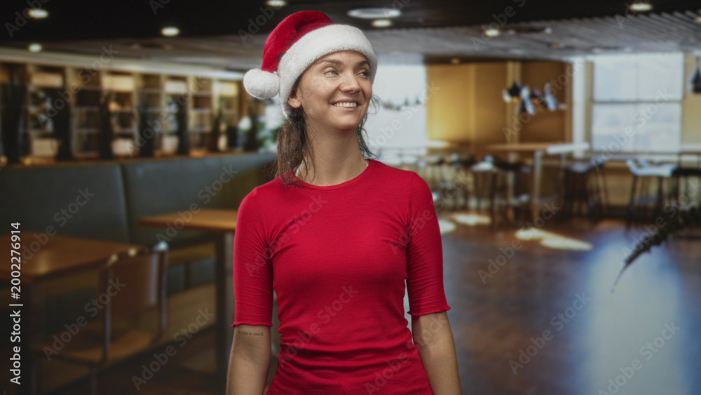 Naklejka premium Woman wearing santa hat smiling and looking up inside a building cafe with wooden tables and chairs; cheerful holiday.