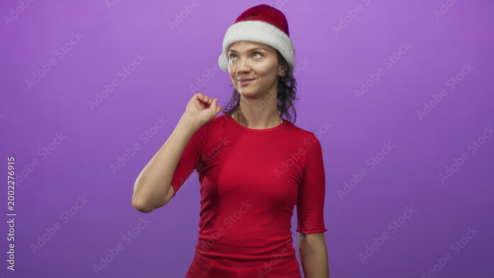 Fototapeta premium Young hispanic woman in santa hat with finger to chin and subtle smirk in purple studio; holiday cheer playful.
