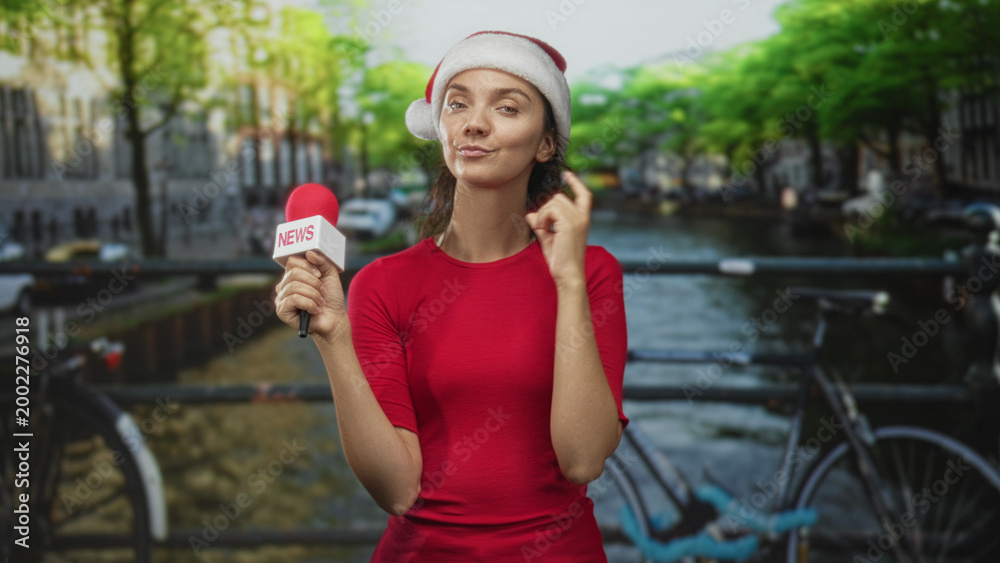 Naklejka premium Young hispanic woman in red shirt and santa hat holds a news microphone while touching her ear on a street canal bridge; festive report.