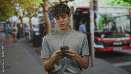 Foto Teen man scrolling a smartphone with hands visible on a city street beside a bus and blurred pedestrians on the sidewalk; quiet contemplation