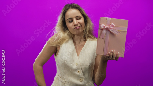Obraz na plátně Woman holding pink gift box with right hand and hand on hip in magenta studio; annoyance disappointment
