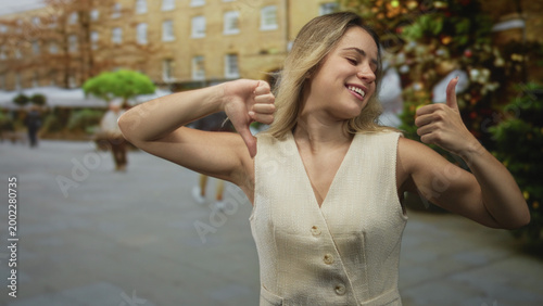 Fotografija Woman showing thumbs up and thumbs down on city street, smiling and grinning while posing in sleeveless vest; ambivalence decision