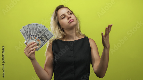 Obraz na plátně Young blonde woman holding a fan of dollar bills with hand raised fanning cash in a green studio; indifference wealth