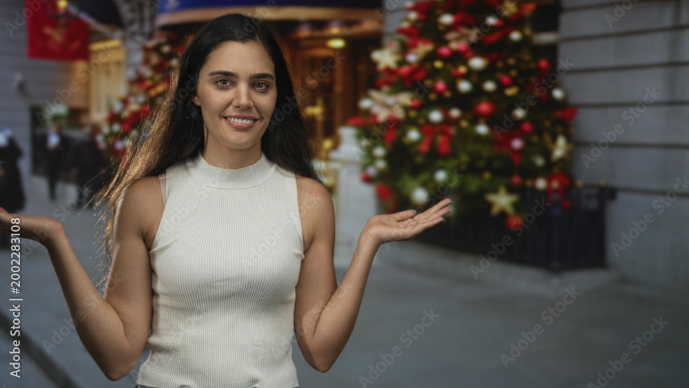 Fototapeta premium Woman in white sleeveless top shows open palms on a city street beside a large decorated christmas tree and storefront entrance; holiday contemplation.