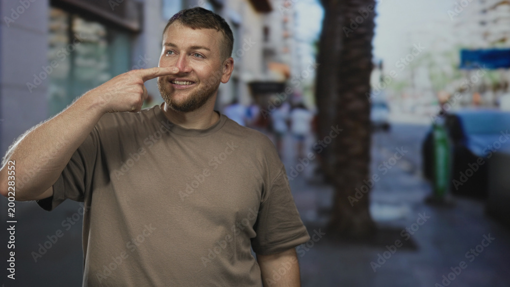 Fototapeta premium Man points finger to nose on a city street, bearded and wearing a brown tshirt, smiling; playful curiosity.