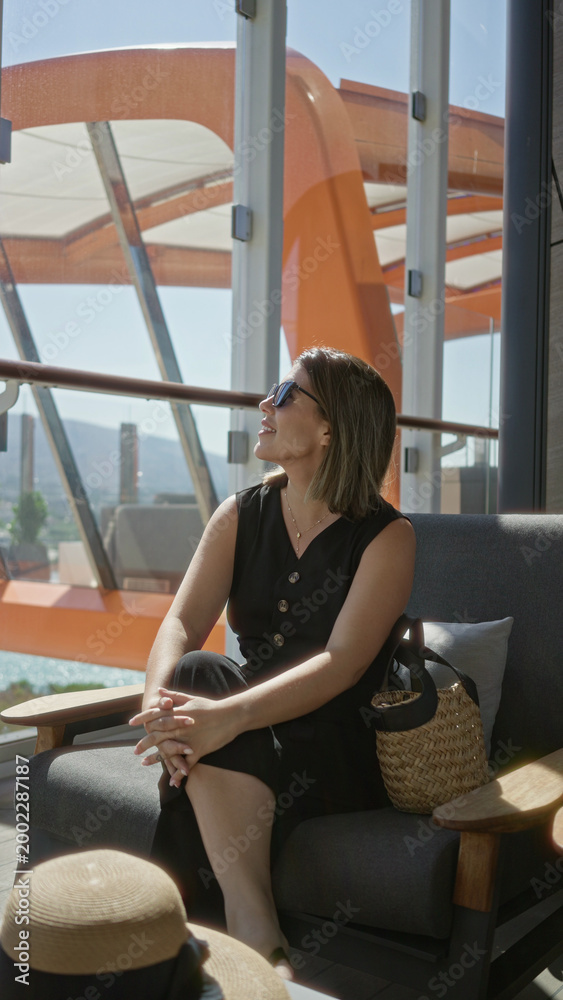 Fototapeta premium Woman holding sunglasses on cruise terrace lounge chair with straw bag and hat beside her while she sits and reaches toward hat; travel leisure calm.