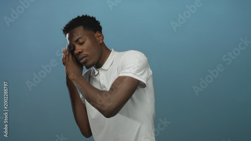 Fotografie Young african american man sleeping with hands at cheek against pale blue background in studio environment; serenity