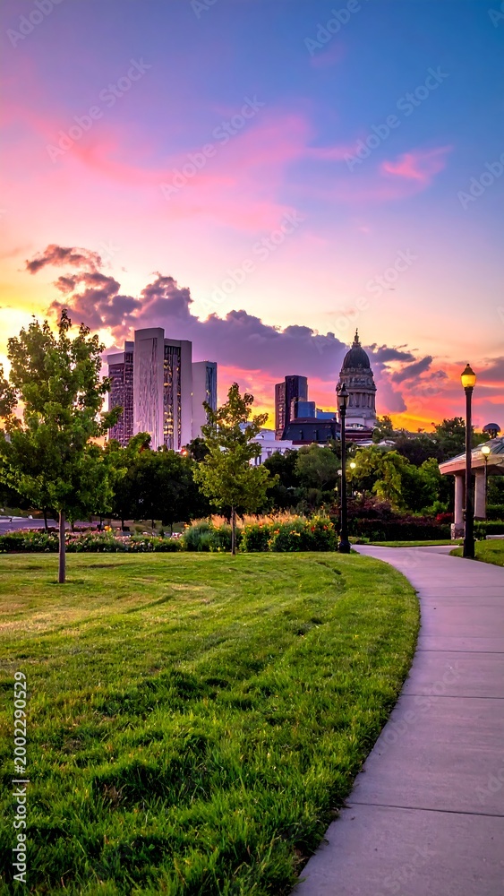 Obraz premium A vibrant sunset casts hues over a cityscape, framed by park greenery and a winding path. Silhouetted buildings reach toward the colorful sky